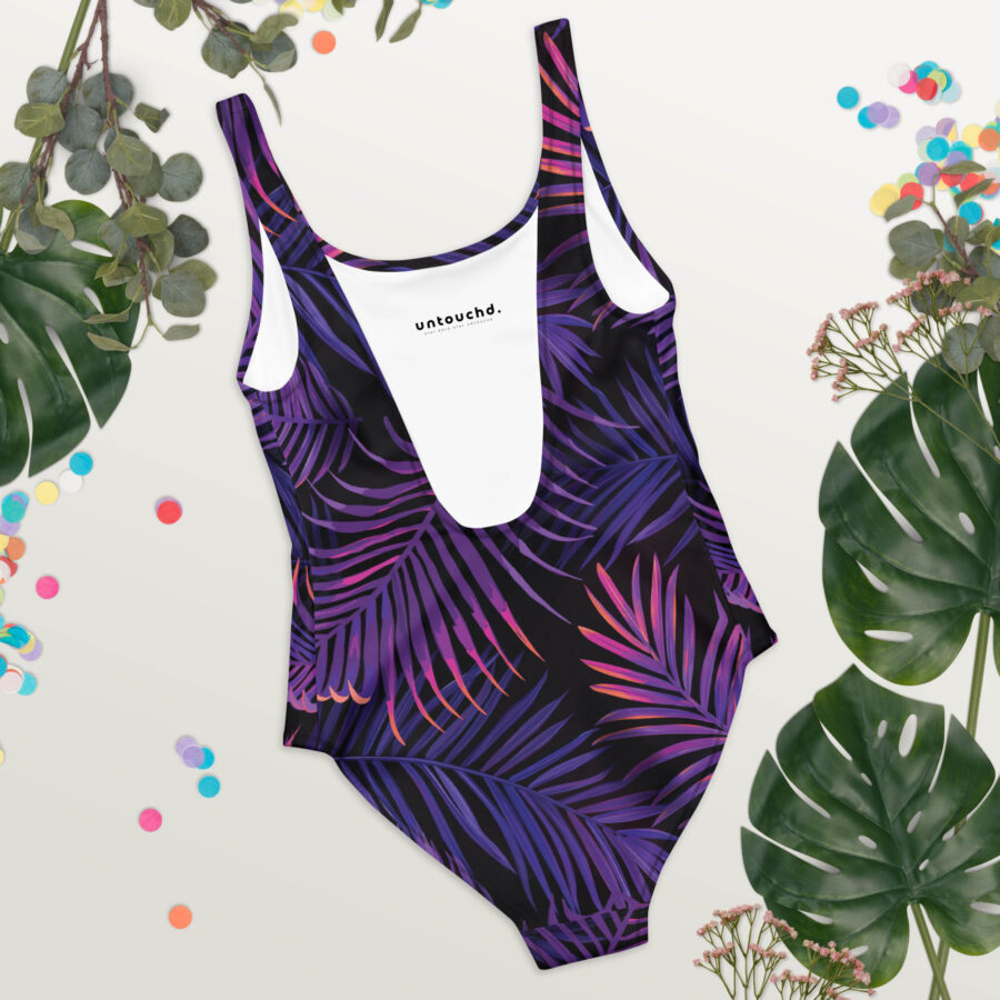 A one-piece swimsuit with a black base and vivid purple and magenta palm leaf pattern, laid flat on a light background surrounded by tropical leaves and colorful confetti. The deep scoop front and white lining reveal the Untouchd logo inside the neckline.