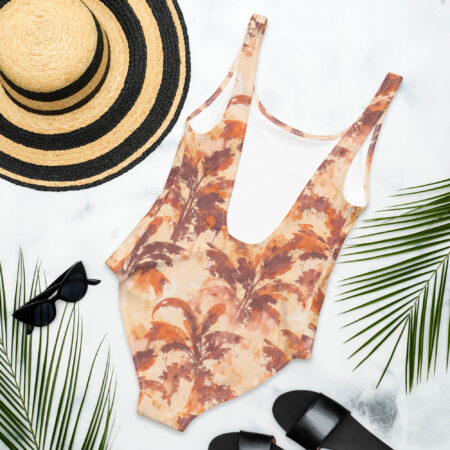 Rustic Autumn Palm One Piece Swimsuit laid flat on a white background with beach accessories including a straw hat, black sunglasses, and sandals — tropical vacation aesthetic.