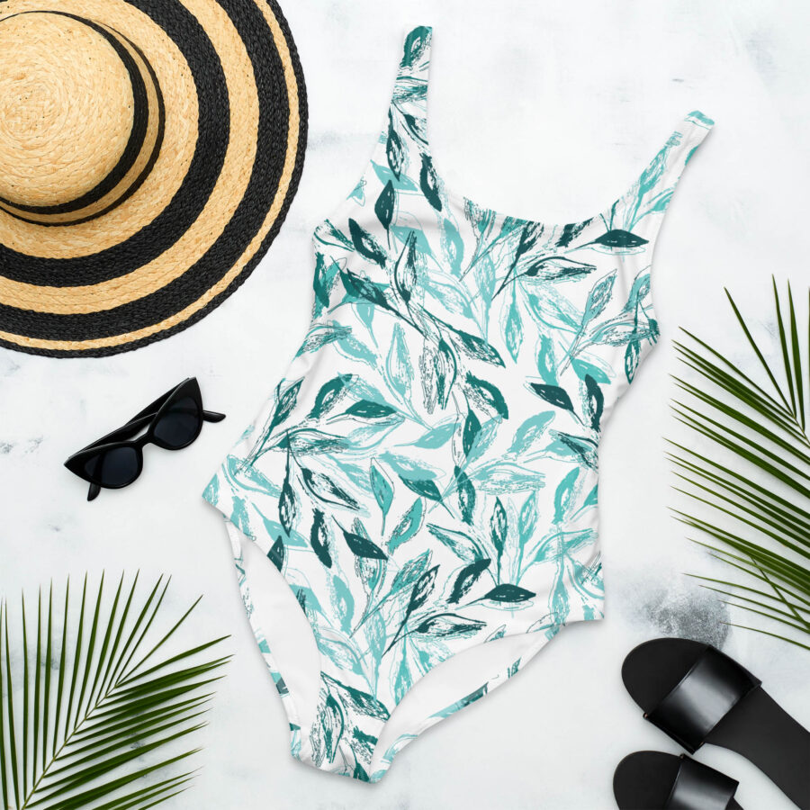 Flat lay of the Botanical Harmony One Piece Swimsuit with teal leaf pattern, paired with a sunhat, black sandals, and sunglasses on a white background.