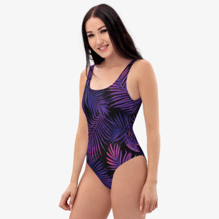 A woman smiles while modeling the Tropic Dusk Palm Leaf Swimsuit in a clean studio setting. The front and side view display the elegant fit and the vibrant purple palm fronds against the dark base.