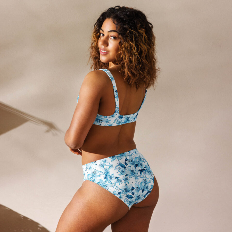 Back view of a woman wearing the Azure Bloom High Waisted Bikini featuring a white base with vibrant blue floral prints, standing in natural sunlight.